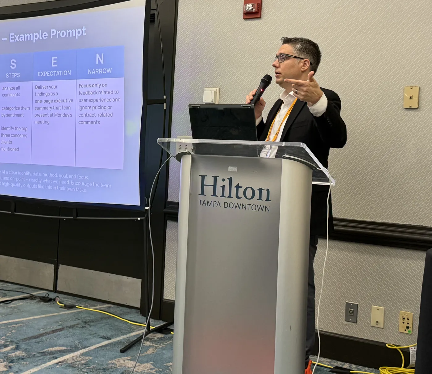 Bryan Dennstedt speaking at a podium beside a slide on prompt writing